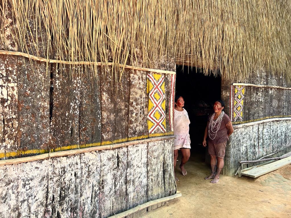 a foto apresenta duas indigenas na maloca no rio negro, elas estão em pé na porta de entrada